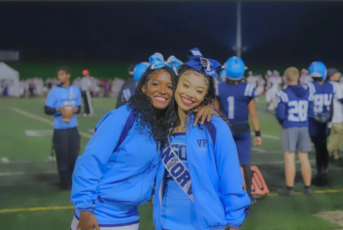 Syrai Smith and a teammate in blue cheer jackets and hair bows, smiling together on the sideline of a night football game.