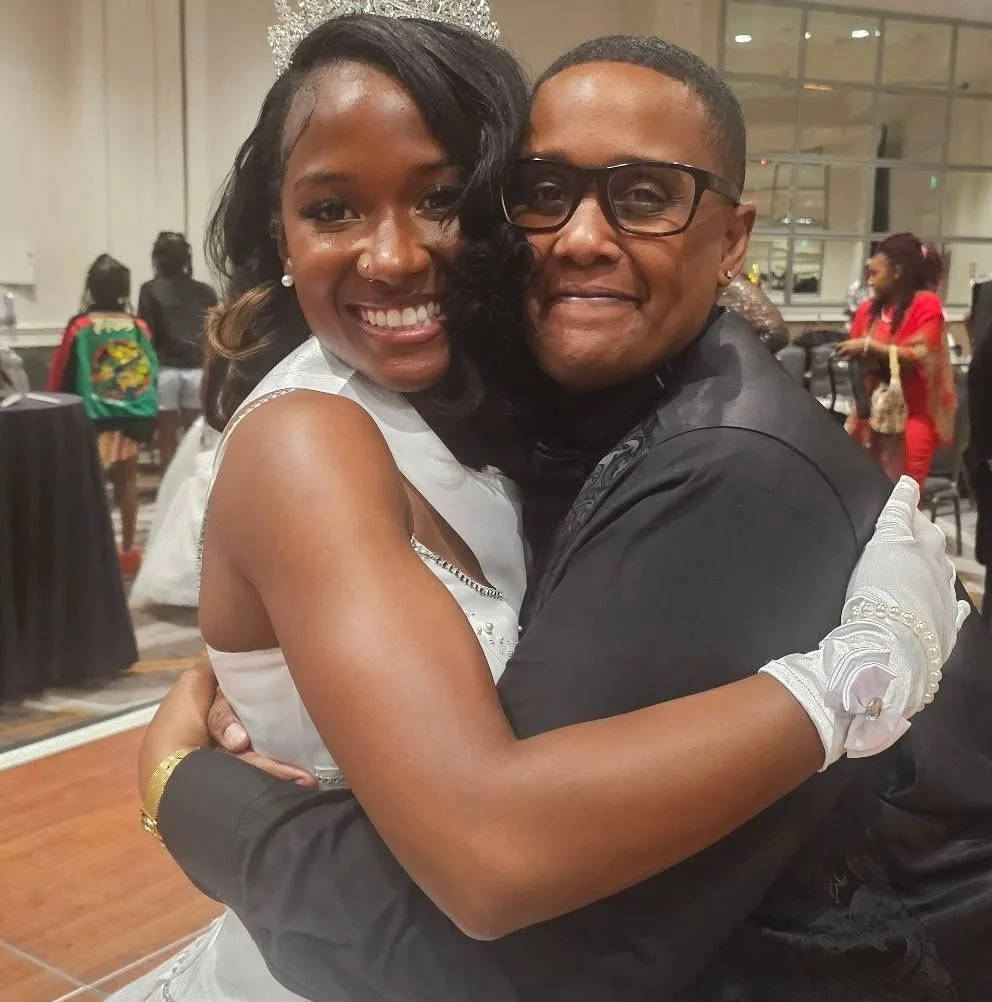 Syrai Smith in a white gown and tiara, smiling as she shares a hug with a loved one.