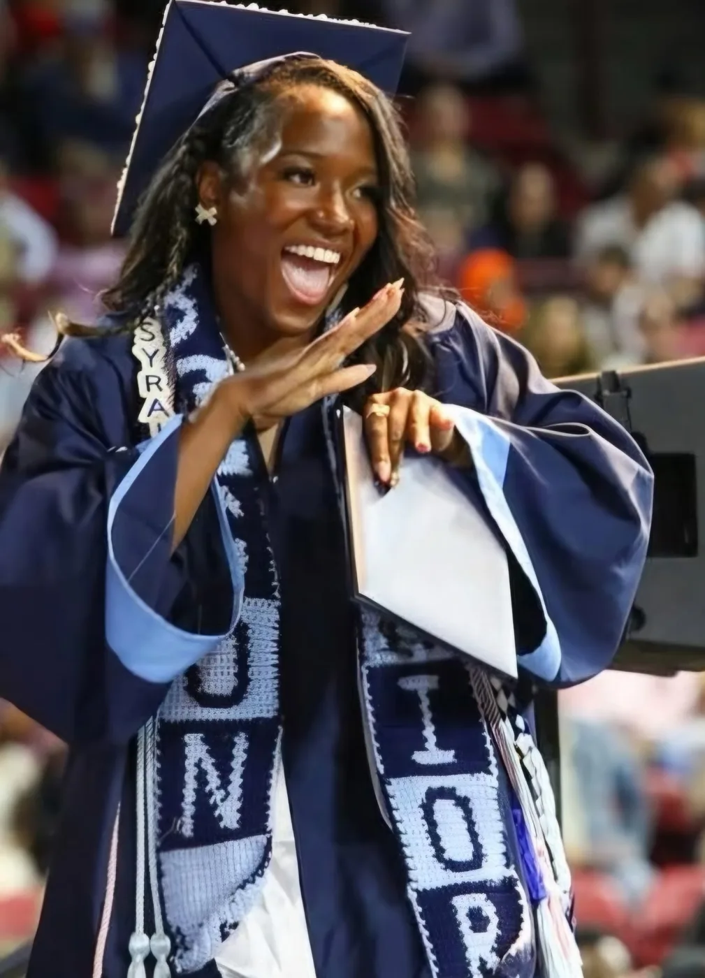 Syrai Smith laughing in a navy graduation cap and gown, holding her diploma.