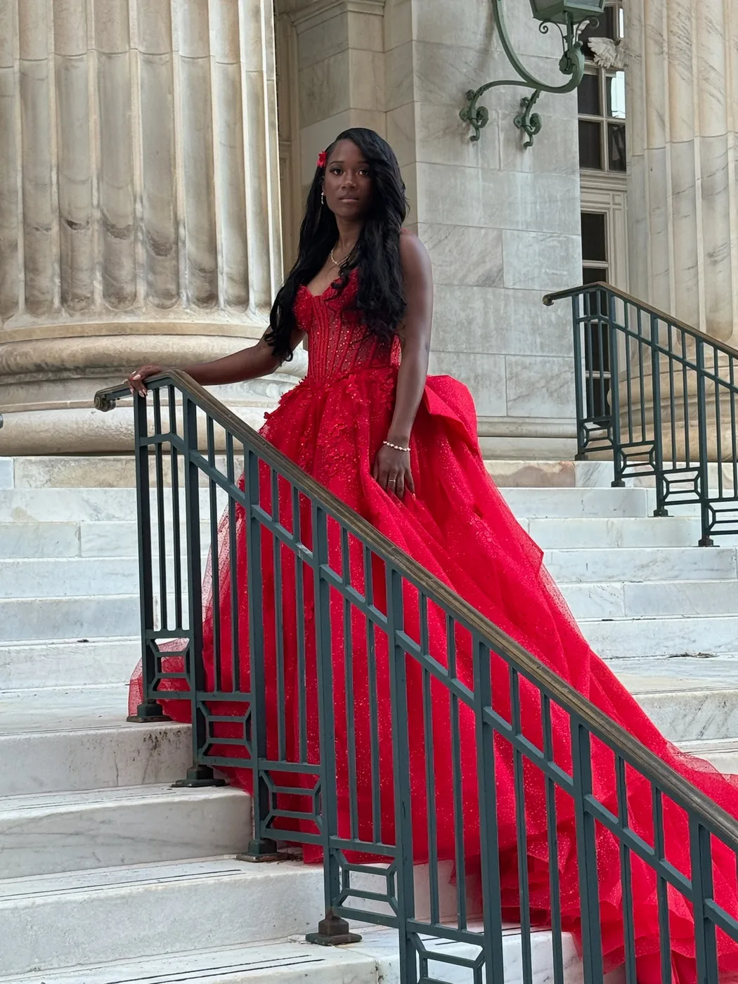 Syrai Smith in a red ball gown on marble steps, one hand resting on an iron stair railing, looking toward the camera.