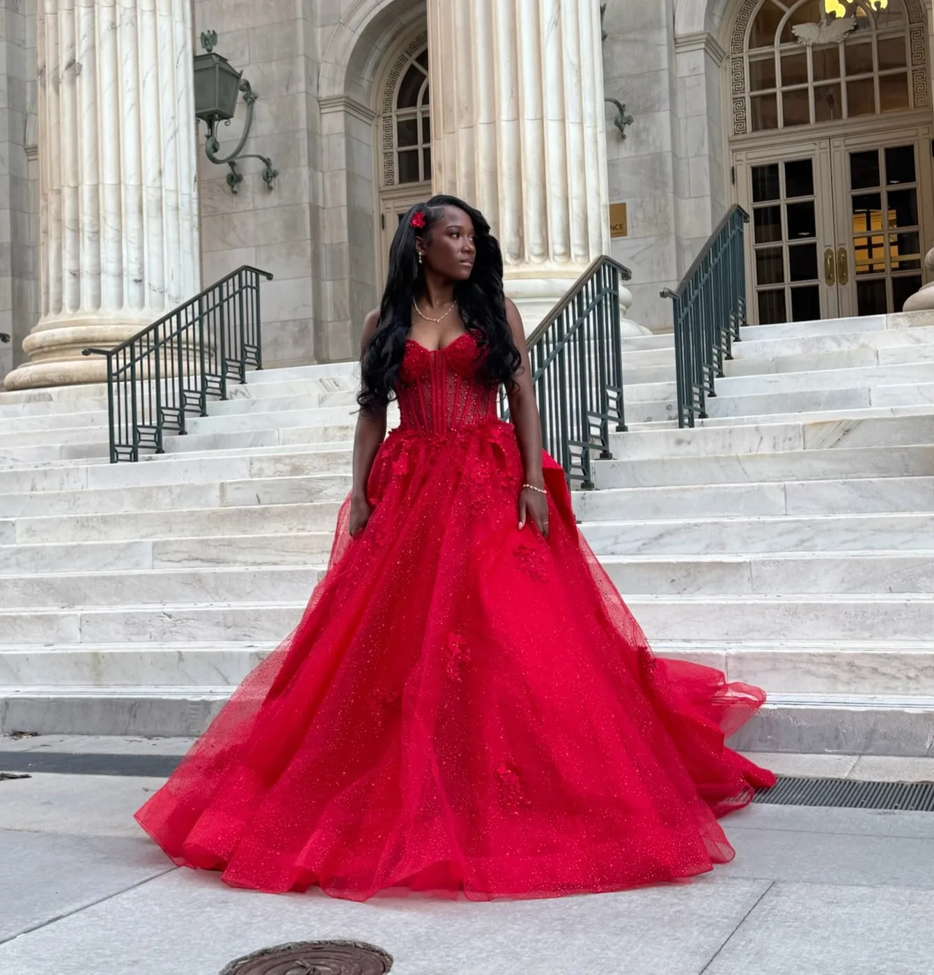 Syrai Smith in a red ball gown, standing on marble steps in front of tall stone columns and looking off to one side.