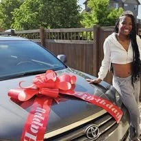 A young woman smiling beside a car topped with a large red bow.